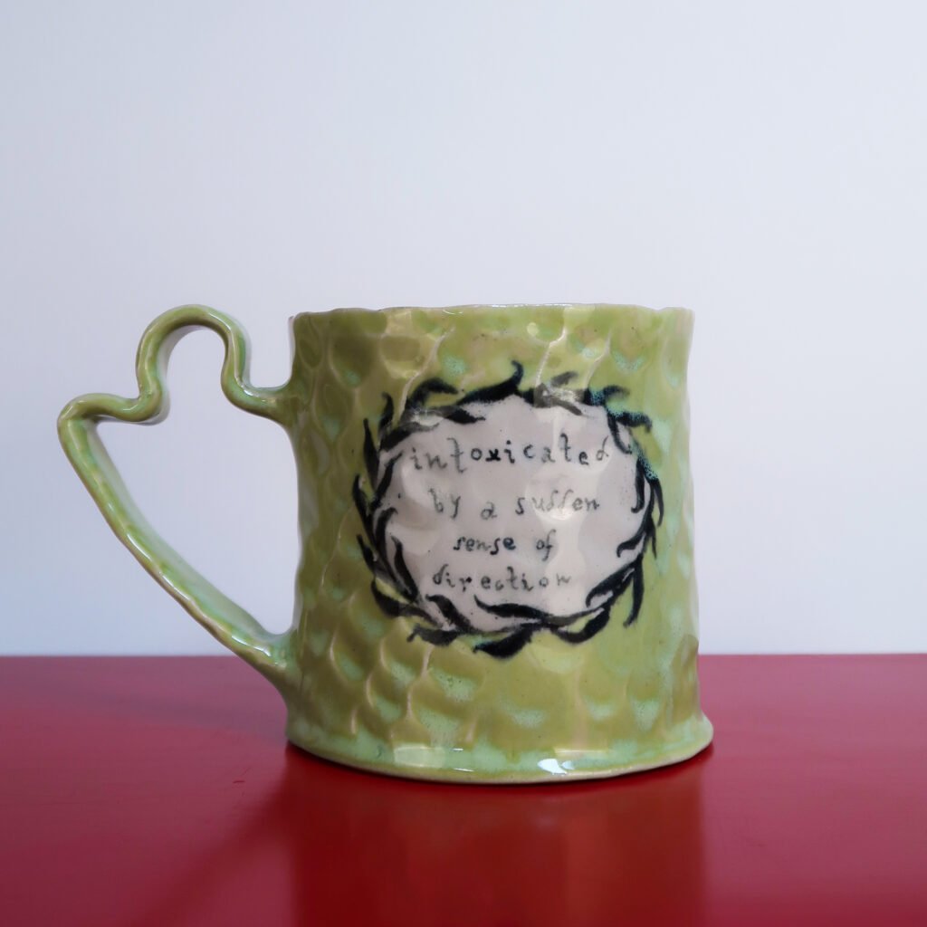handmade green ceramic mug with a fancy handle, fingerprint texture, the words intoxicated by a sudden sense of direction