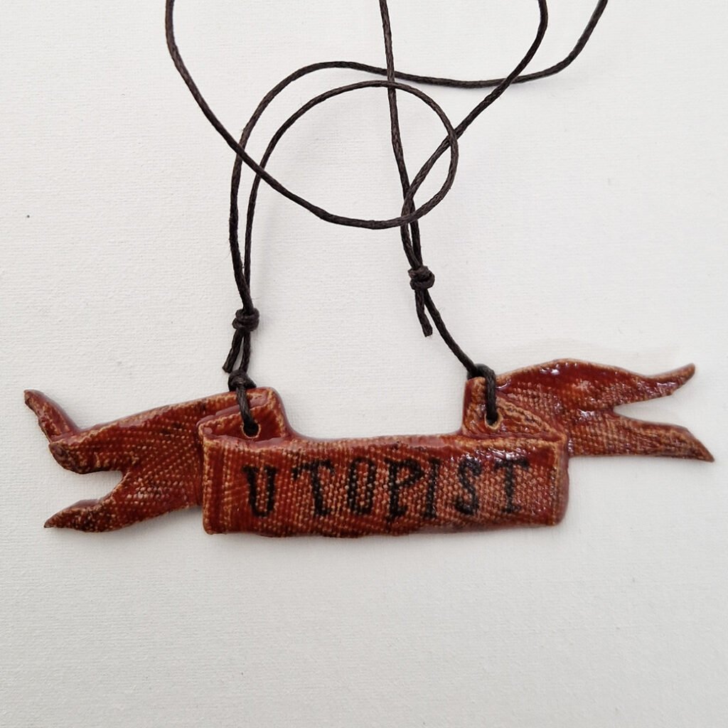 a ceramic pendant in the shape of a tattered flag with the word utopist