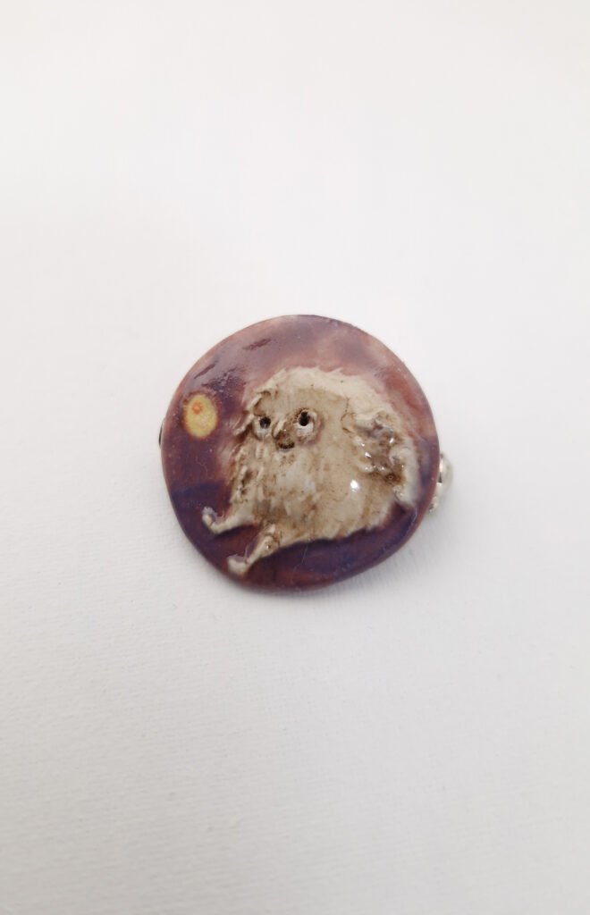small round ceramic brooch with a small silly feathered character sitting down
