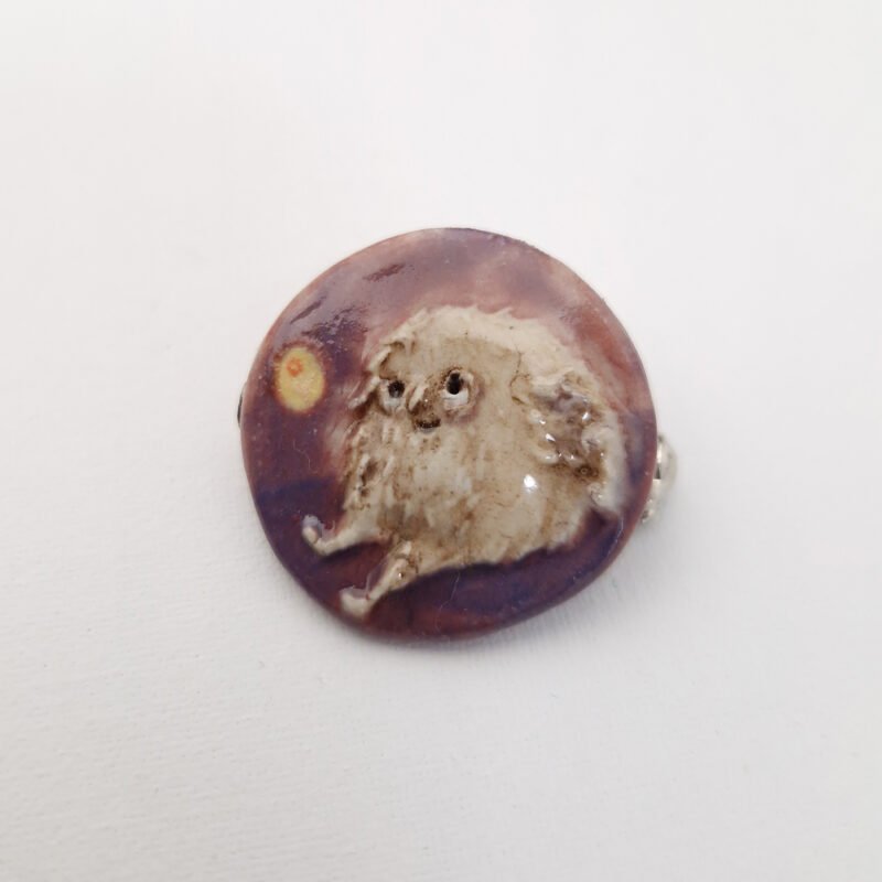 small round ceramic brooch with a small silly feathered character sitting down