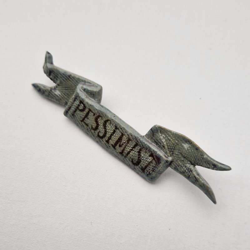 ceramic brooch in the shape of a tattered flag with the word "pessimist" drawn on