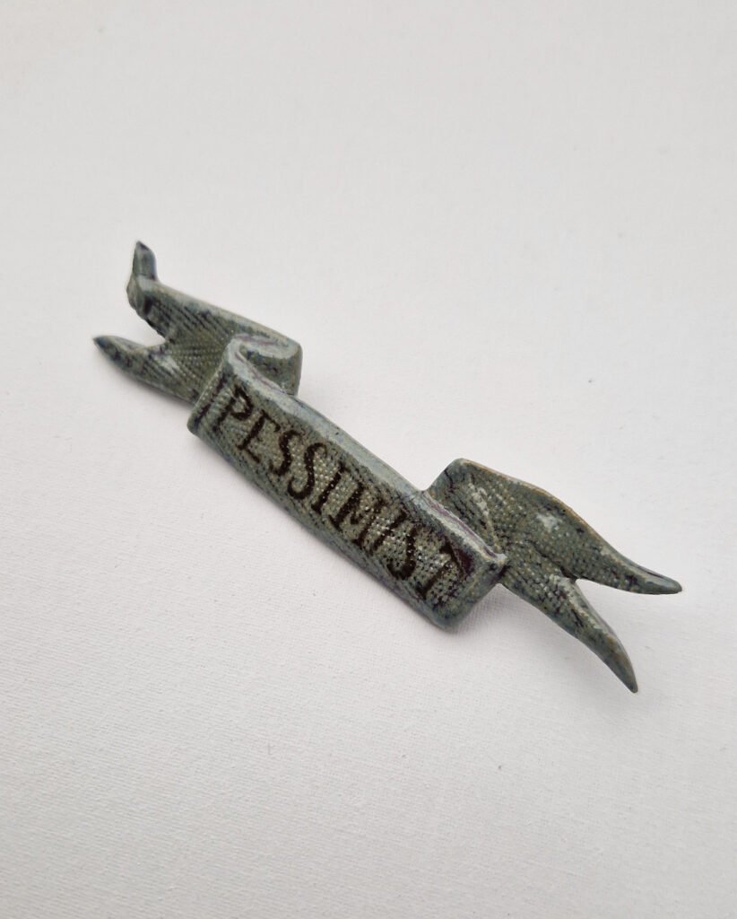 ceramic brooch in the shape of a tattered flag with the word "pessimist" drawn on